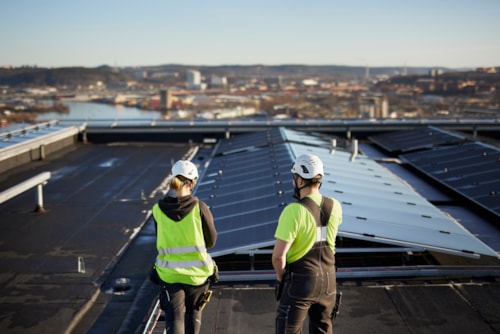 Two workers with bright yellow safety vests and helmets seen from behind looking at solar panels installed on the roof of a building